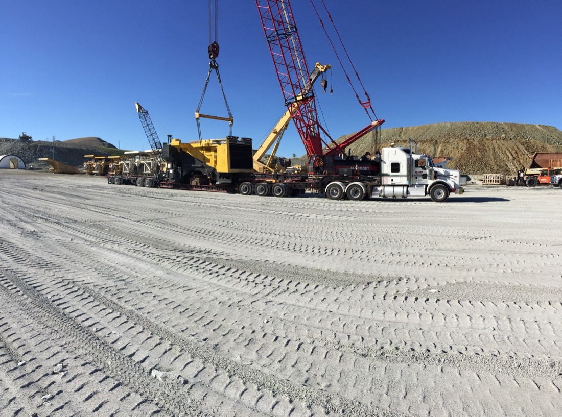 Yellow mining machinery on multi-axle trailer at mine site, lattice crawler crane in background