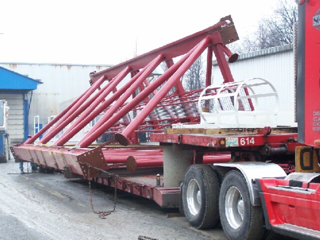 Red lattice mast (drill rig / pile-driver boom) with operator cage on low-bed trailer