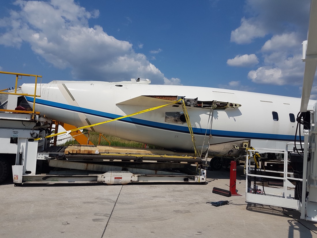 Commercial aircraft fuselage (Boeing-style narrow-body, white with blue stripe) on specialized aerospace transport dollies