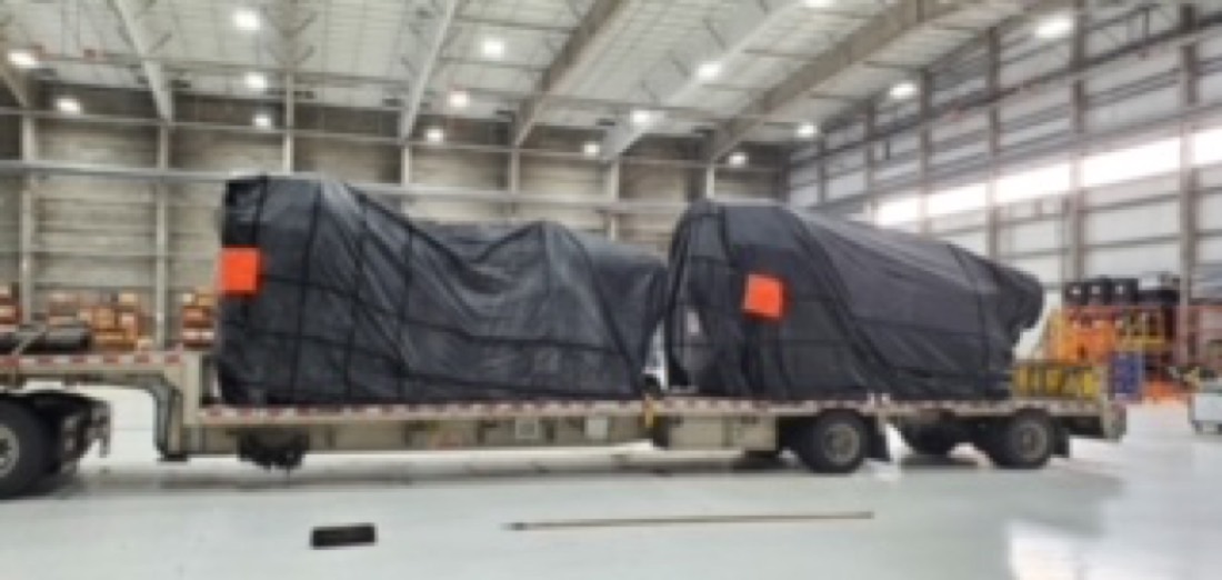 Two tarp-covered large cargo items (likely helicopter fuselages or aircraft components) on step-deck flatbed