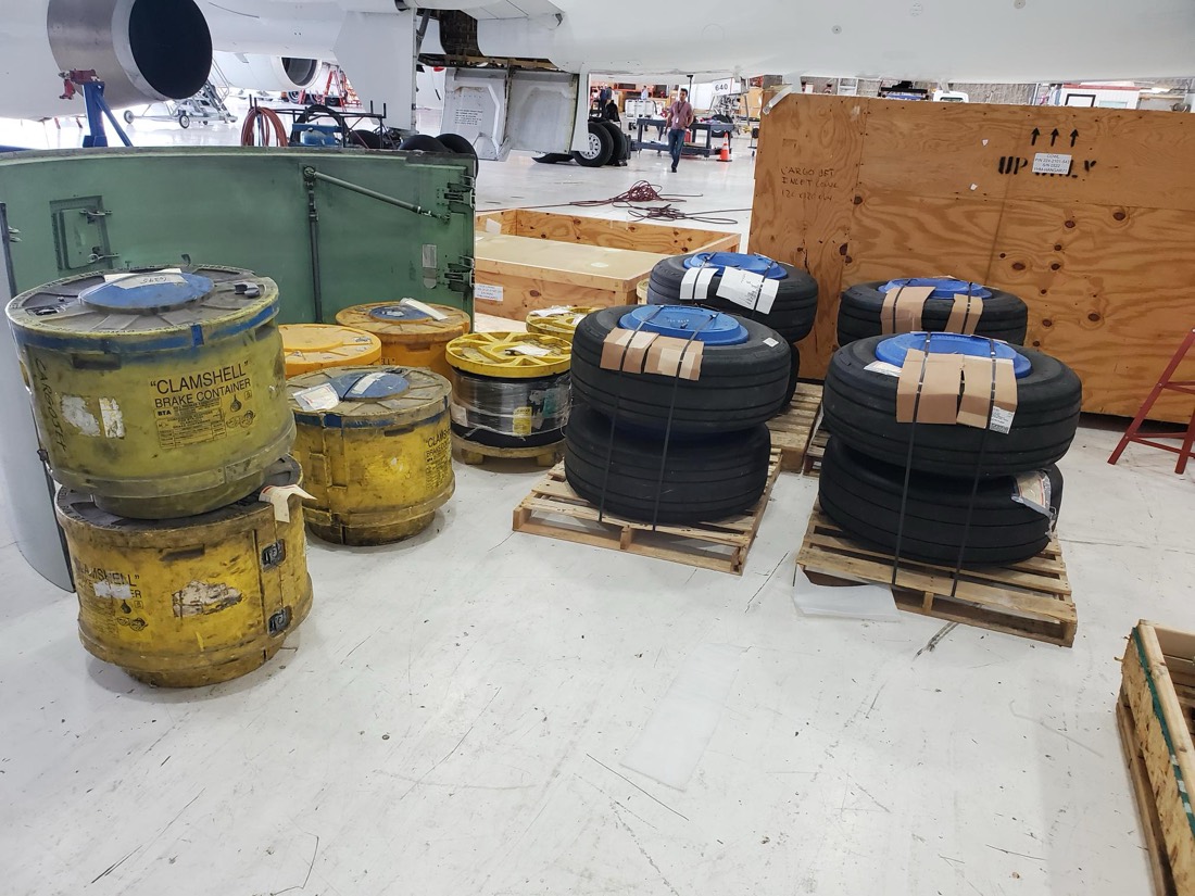 Aircraft tires on pallets plus yellow CLAMSHELL drums (engine-cowl containers) staged in hangar