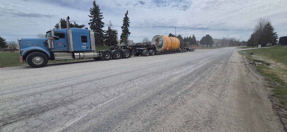 Same yellow TBM drum loaded on modular trailer, blue Kenworth tractor — full rig on road