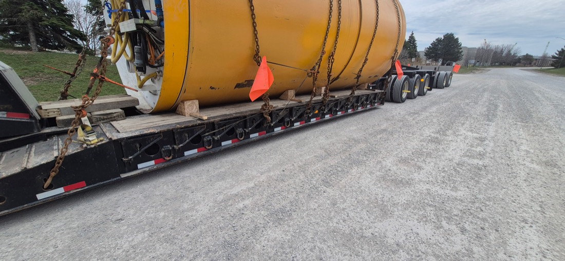Close-up of yellow TBM drum chain-secured on modular trailer (oversize flags visible)