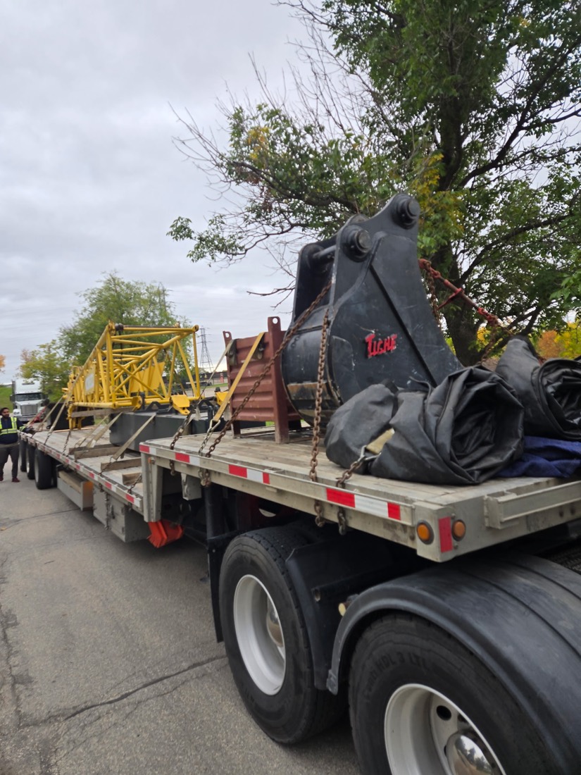 Large black excavator bucket on flatbed; yellow crane lattice boom section on adjacent trailer