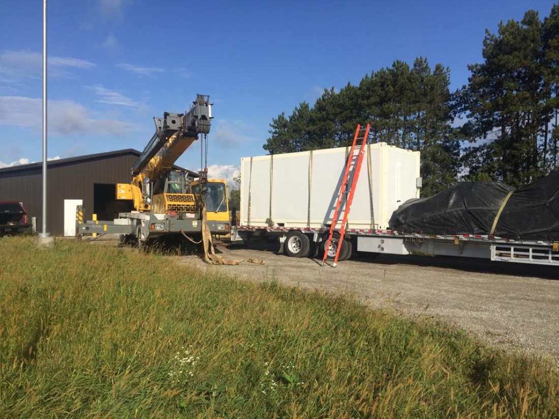 Yellow Grove mobile crane next to flatbed with white modular shelter / portable enclosure; tarped load on adjacent trailer