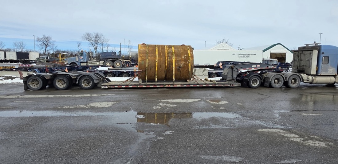 Rust-yellow steel drum / mill section on multi-axle lowboy at Heavy Lift Cargo Solutions yard (winter)