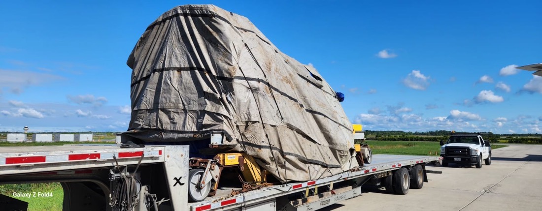 Tarp-wrapped aerospace component (jet engine on stand) on step-deck flatbed at airfield