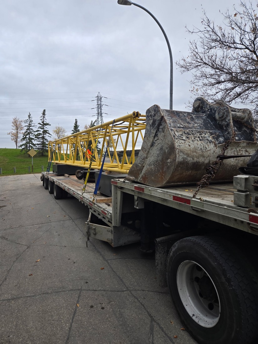 Yellow crane lattice boom sections on flatbed, large excavator bucket on second trailer