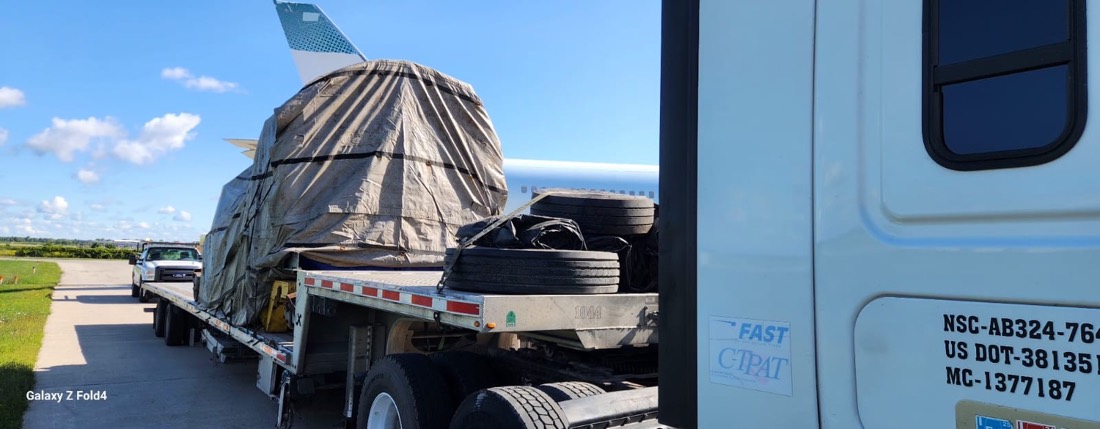 Tarp-wrapped jet engine on stand + aircraft tires on step-deck at airfield (Bombardier-style aircraft behind)