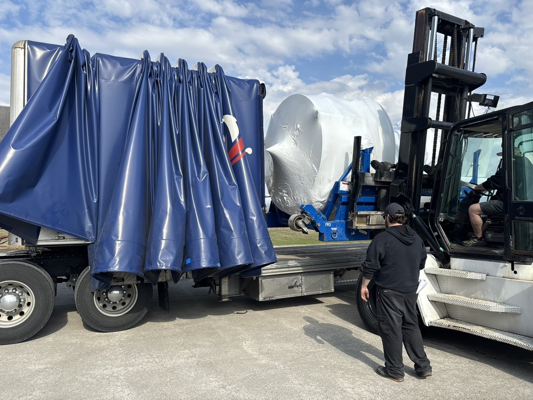 White shrink-wrapped pressure vessel being loaded into blue Conestoga rolling-tarp trailer by forklift