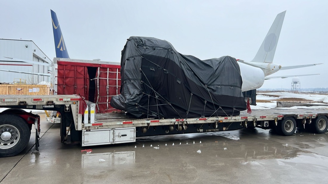 Black-tarped aircraft engine on stand on step-deck flatbed at airport (Boeing aircraft behind)
