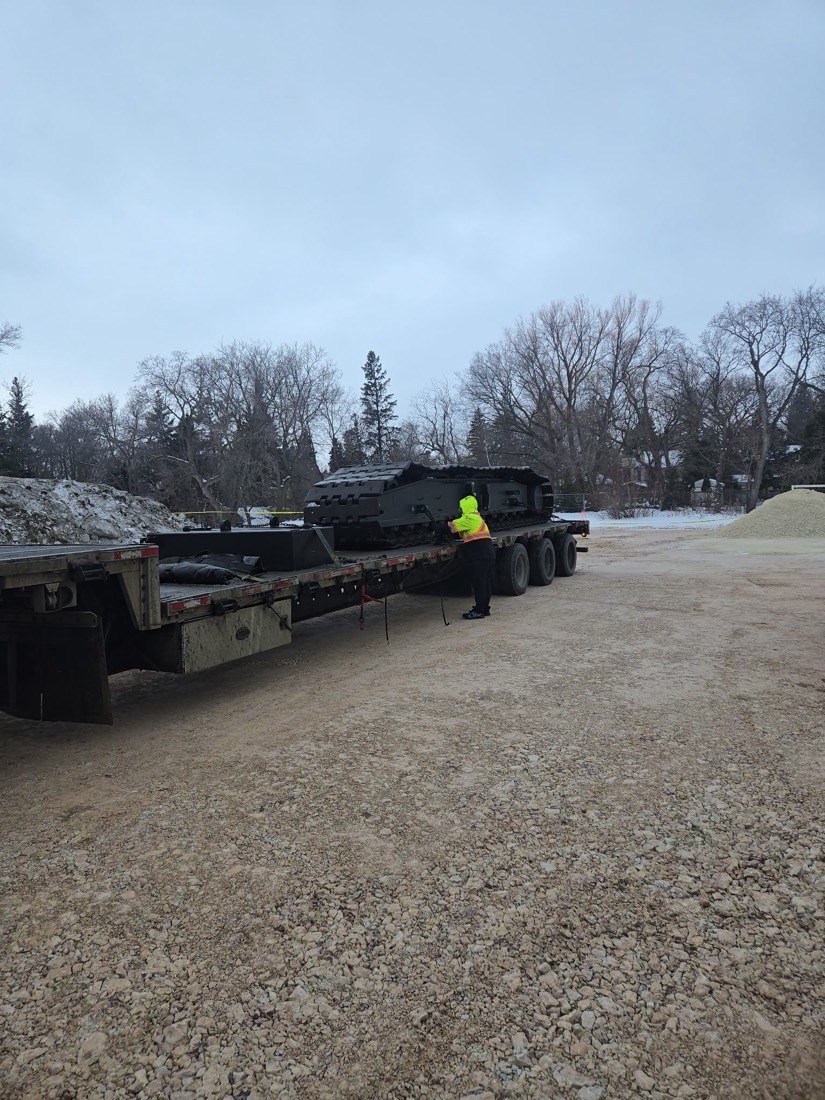 Worker beside near-empty lowboy trailer with small dark cargo piece at rear (winter yard)