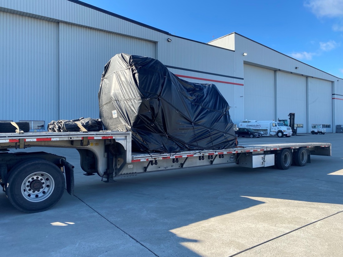 Black-tarped Cargojet jet engine on stand on step-deck flatbed beside Cargojet hangar
