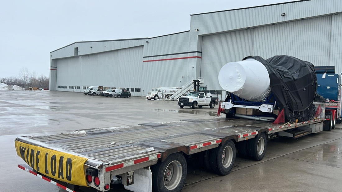 Jet engine on stand (white shrink-wrap + black tarp) on WIDE LOAD step-deck at Cargojet hangar