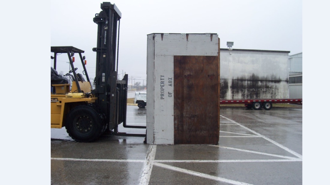 Yellow forklift moving large wooden shipping crate, tanker trailer in background