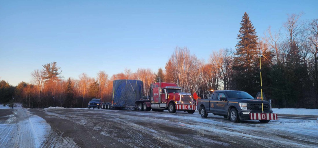 Red Peterbilt with blue-tarped oversize load on step-deck, escorted by F-150 pilot vehicle in winter