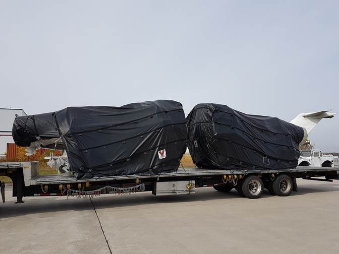 Two tarp-wrapped aerospace components (jet engines on stands) on step-deck flatbed at airfield
