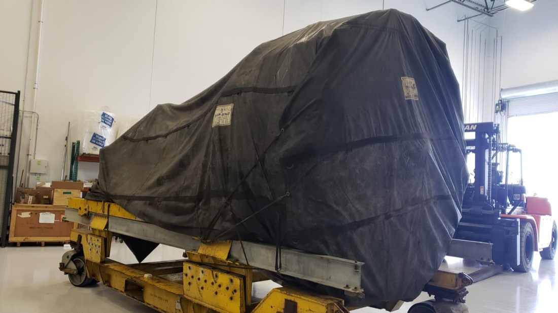 Same tarp-wrapped jet engine on yellow stand inside MRO facility — wider view with forklift