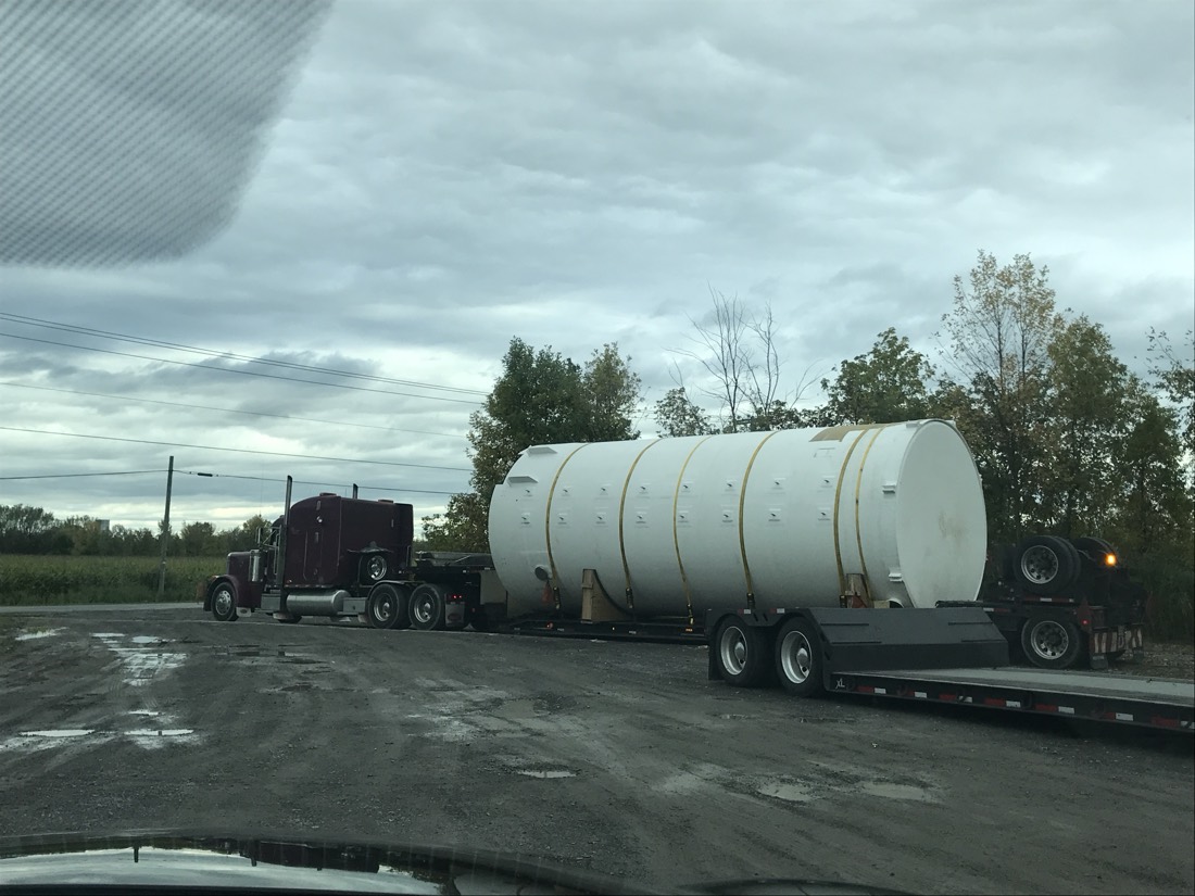 Same large fiberglass storage tank on extendible flatbed pulled by maroon Peterbilt