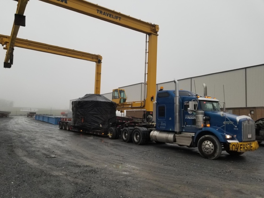 Blue Kenworth with tarp-wrapped oversize load under TRAVELIFT gantry crane at fab yard