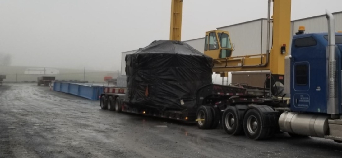 Blue Kenworth with tarp-wrapped oversize cargo on lowboy beside yellow gantry crane (foggy)