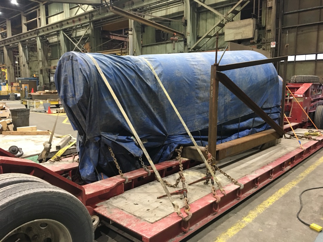 Blue-tarp-wrapped cylindrical cargo (mill roller / shaft) on red lowboy in fab shop