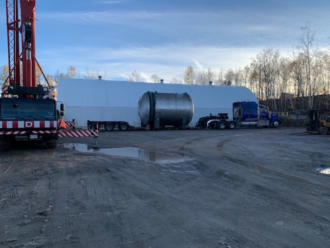 White insulated vessel + stainless pressure vessel on flatbeds, blue Peterbilt and Liebherr crane at job site
