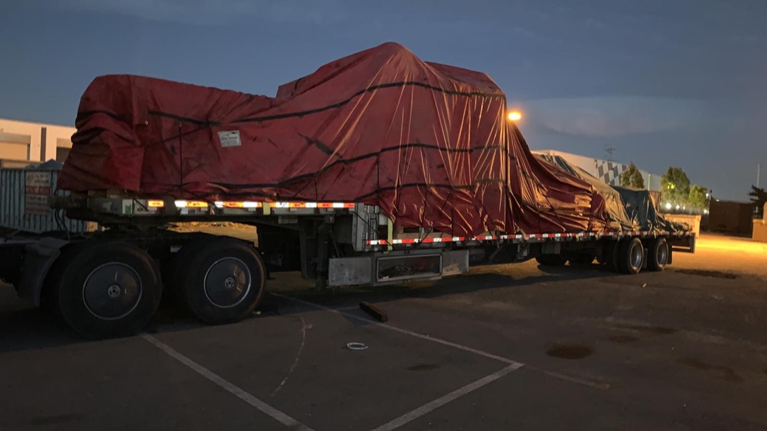 Red-tarped oversize machinery on flatbed at dusk parking lot