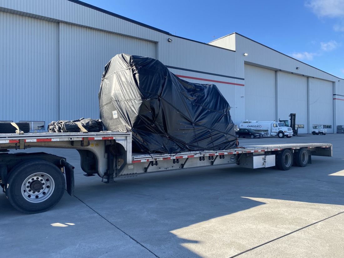 Black-tarped jet engine on stand on step-deck flatbed at Cargojet hangar