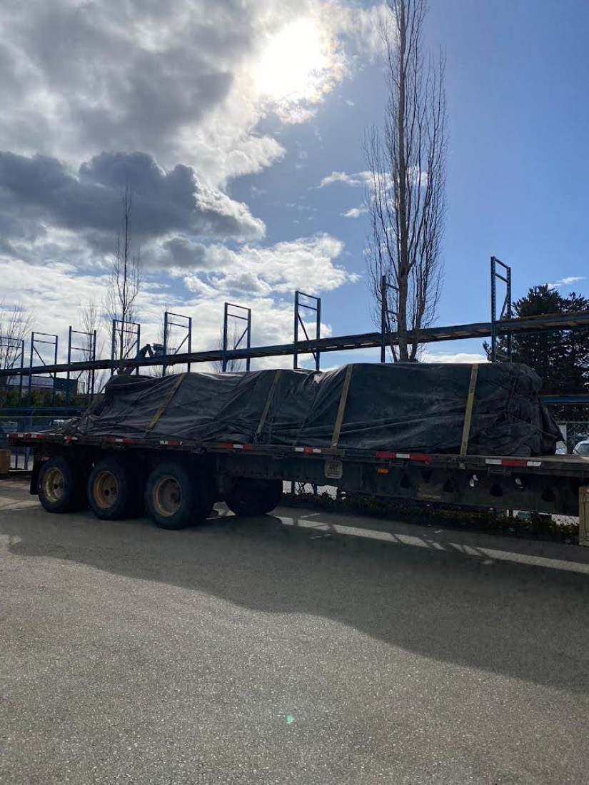 Black-tarp-covered industrial cargo on tri-axle flatbed at storage yard with empty racking