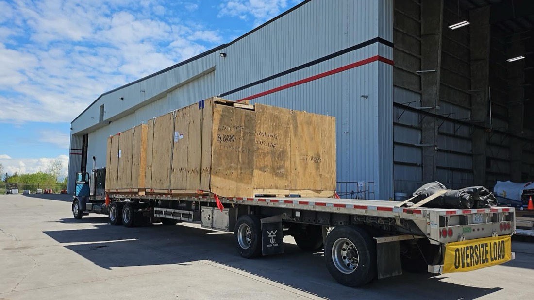 Long aerospace shipping crate on OVERSIZE LOAD step-deck flatbed beside hangar (duplicate of cj od.jpg)