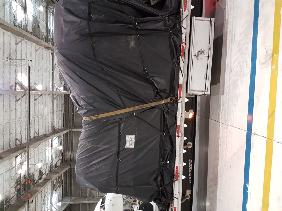 Black-tarp-wrapped cargo on flatbed inside hangar/warehouse (Kinedyne strap)