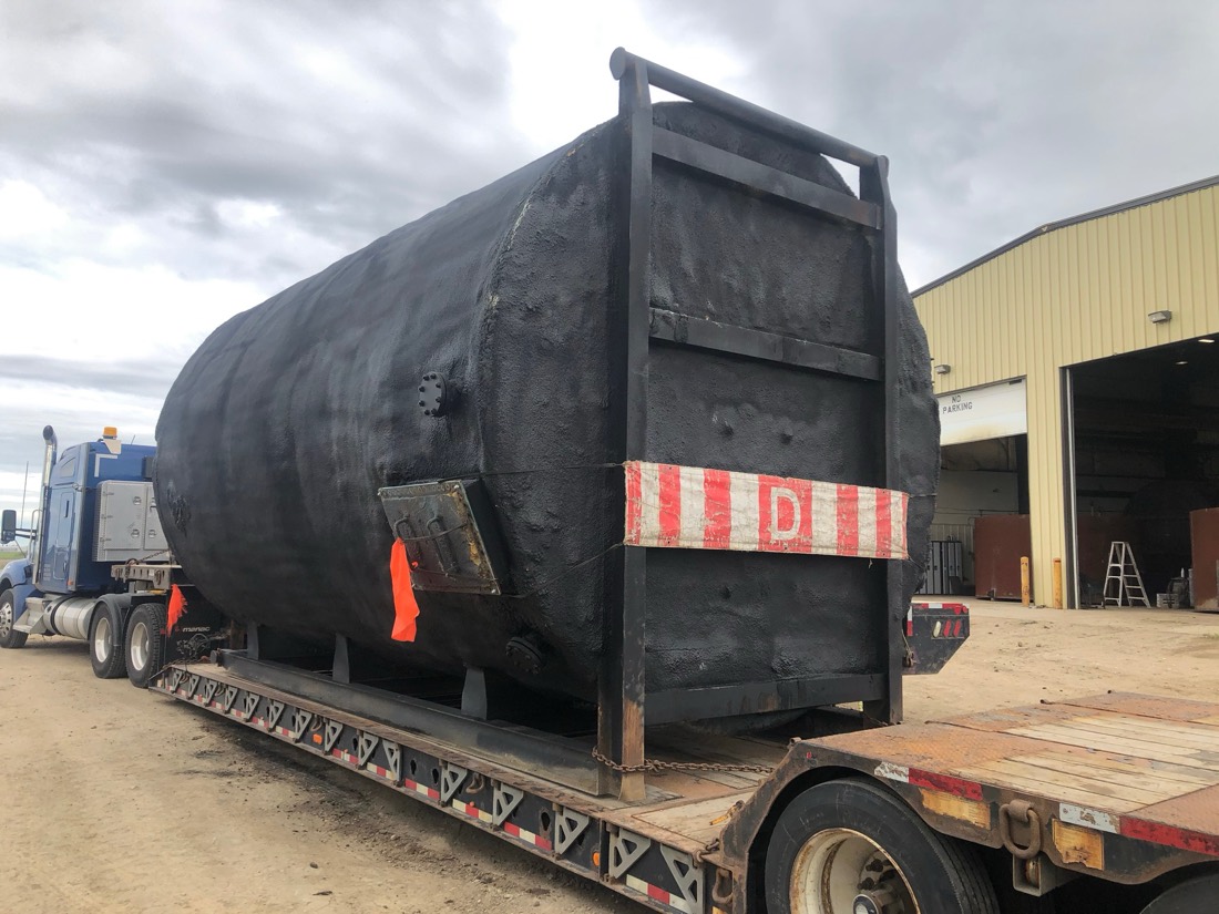 Same black-coated insulated tank on lowboy — side view at industrial yard