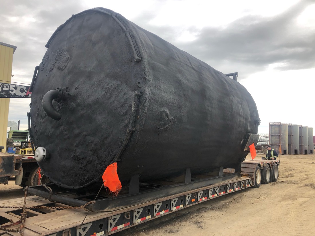 Same black-coated insulated tank on lowboy — opposite end view at industrial yard