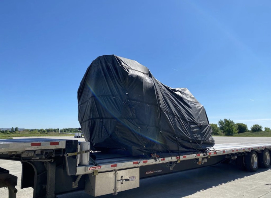 Same GE jet engine on stand black-tarp-wrapped on Great Dane step-deck flatbed at airfield