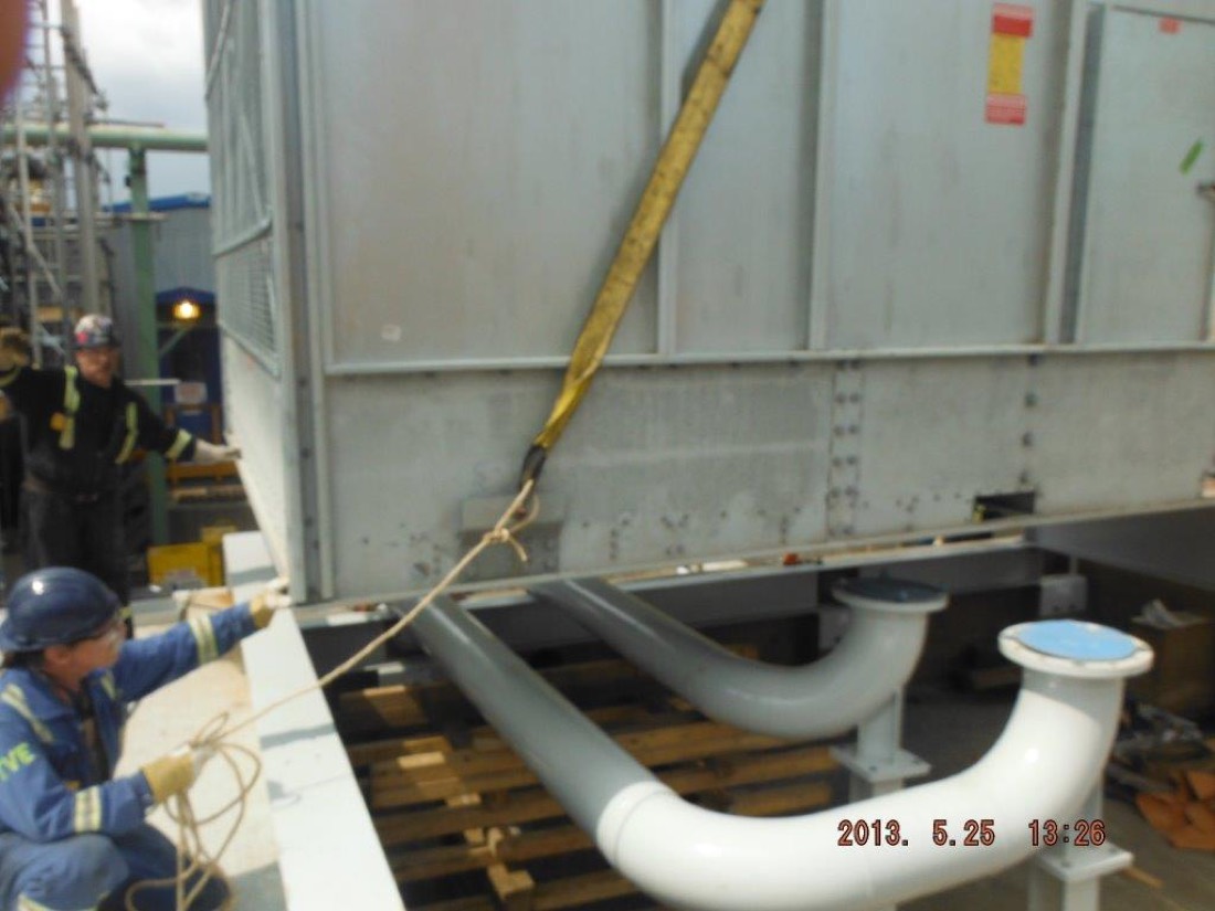Workers positioning cooling tower module above white piping manifolds at refinery