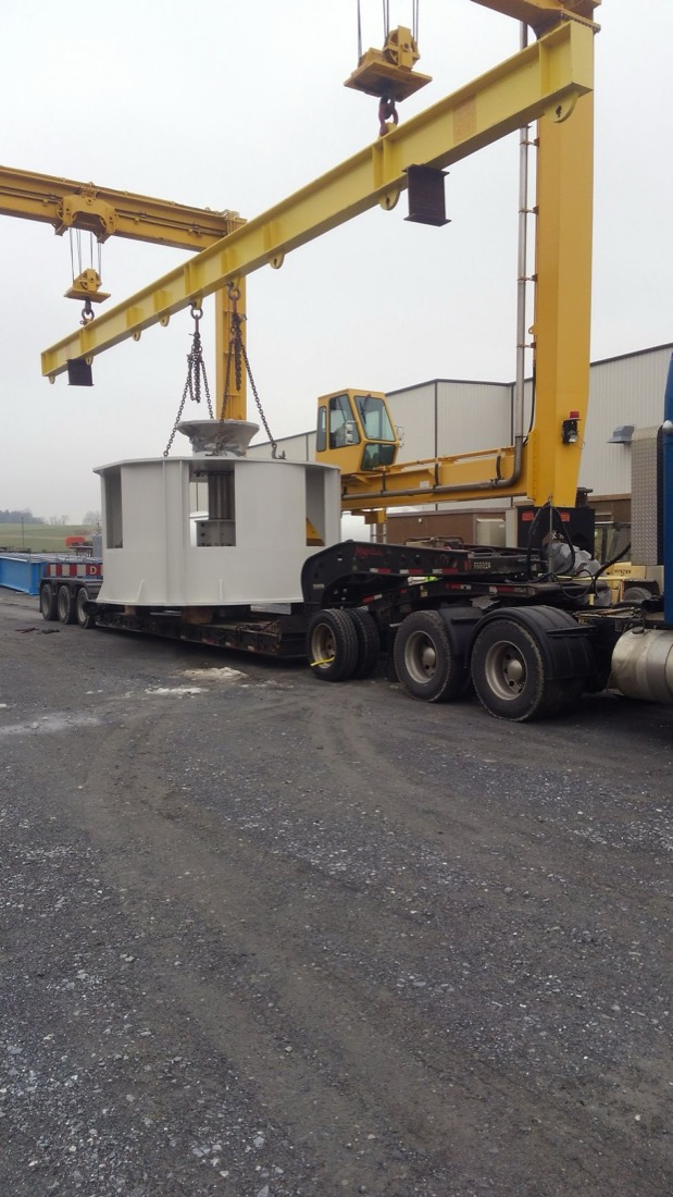 White industrial enclosure rigged under overhead gantry crane onto lowboy