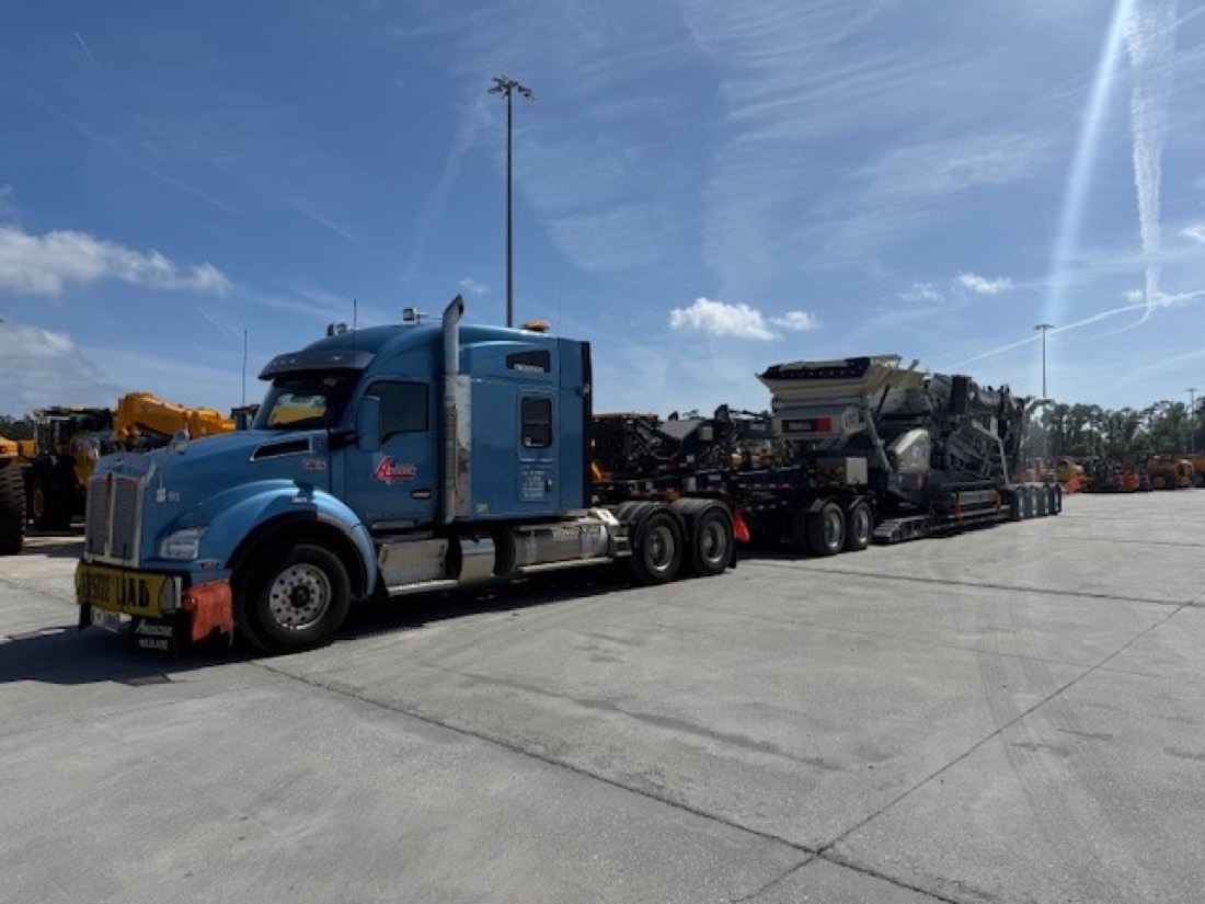Blue Kenworth pulling tracked Metso crusher on lowboy across equipment yard