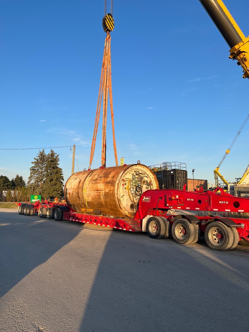 Tunnel boring machine cutterhead suspended in slings above red multi-axle trailer