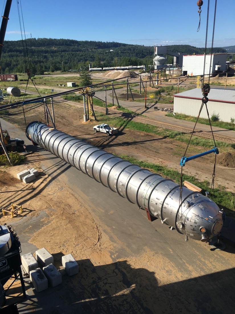Long horizontal stainless distillation column being airlifted by twin cranes at refinery yard