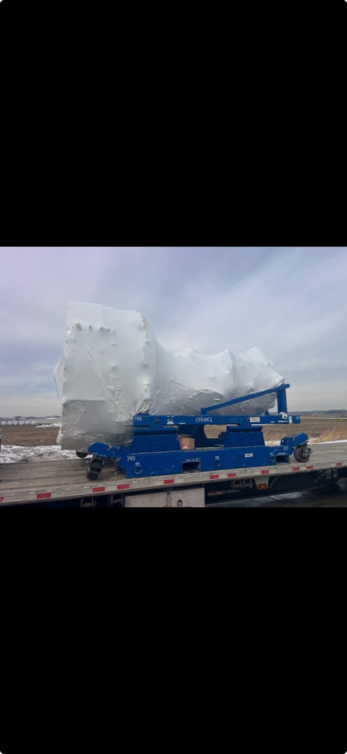 Shrink-wrapped angular component nested on blue beam trailer cradle, end view
