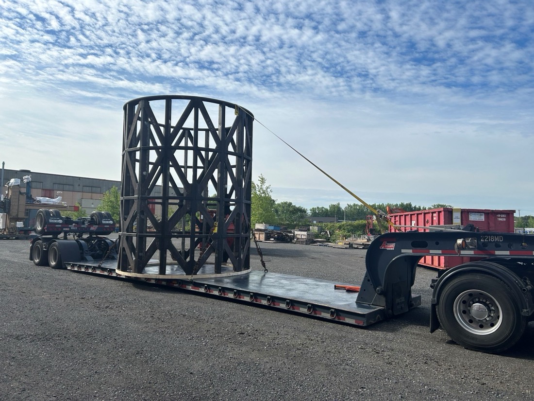 Bare black trommel cage strapped to stepdeck at industrial yard with summer trees behind