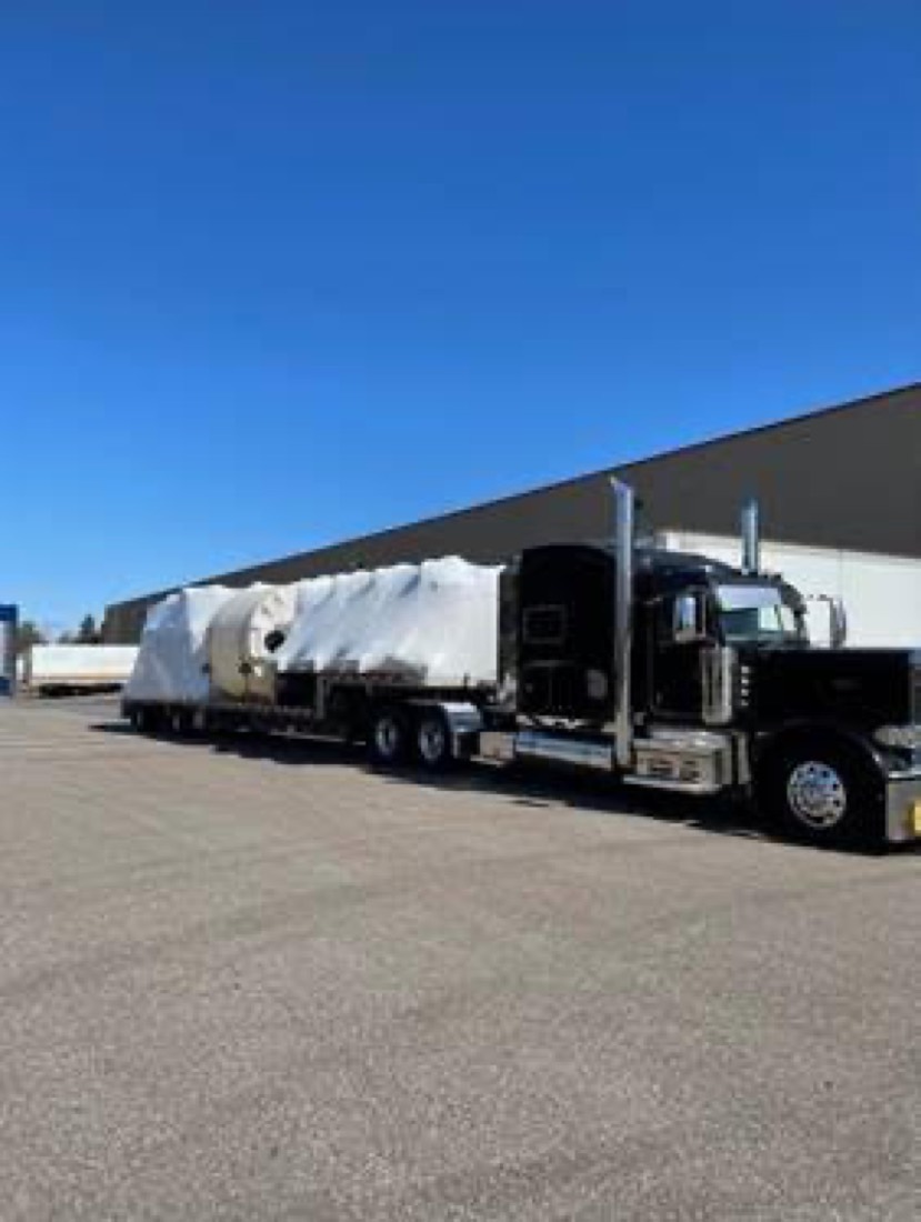 Black Peterbilt with white shrink-wrapped aircraft component on flatdeck outside warehouse