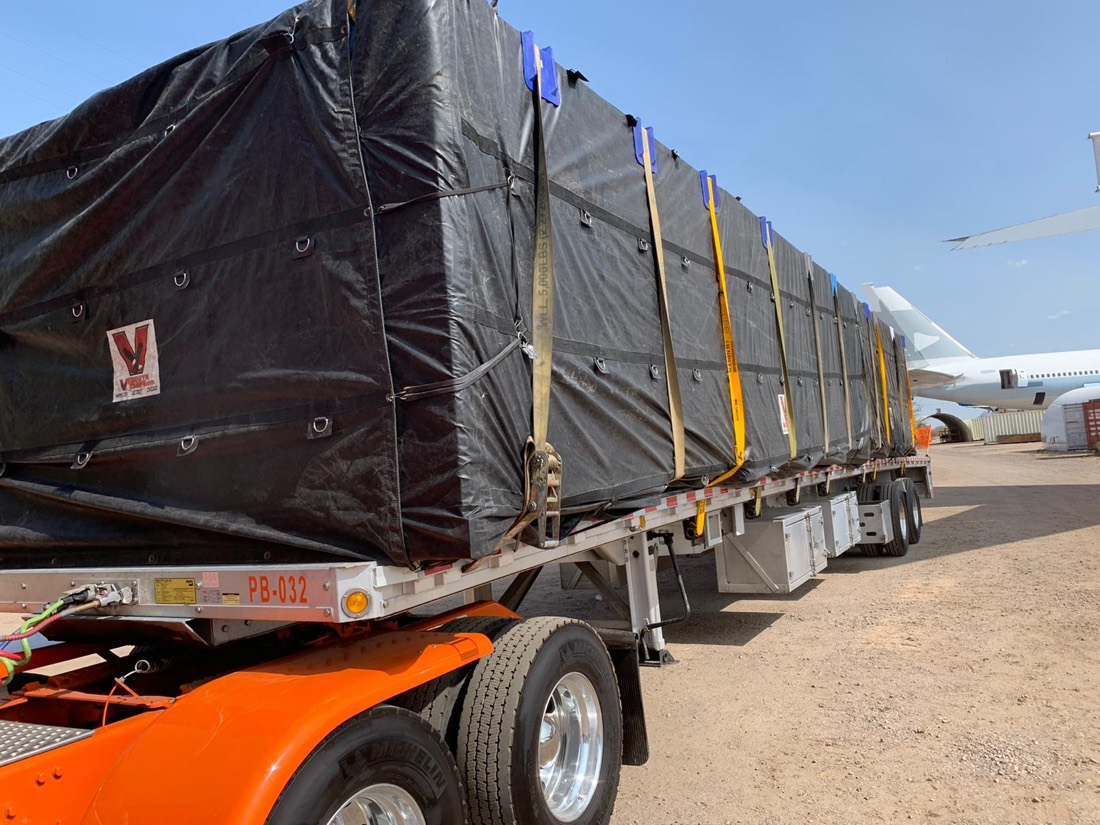 Black-tarped V-marked equipment crate tied down to flatdeck beside parked airliner at desert ramp