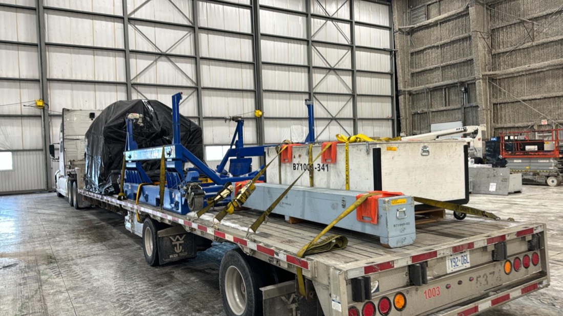 Long crate B71006-341 strapped beside aero cradle and tarped load on stepdeck inside hangar