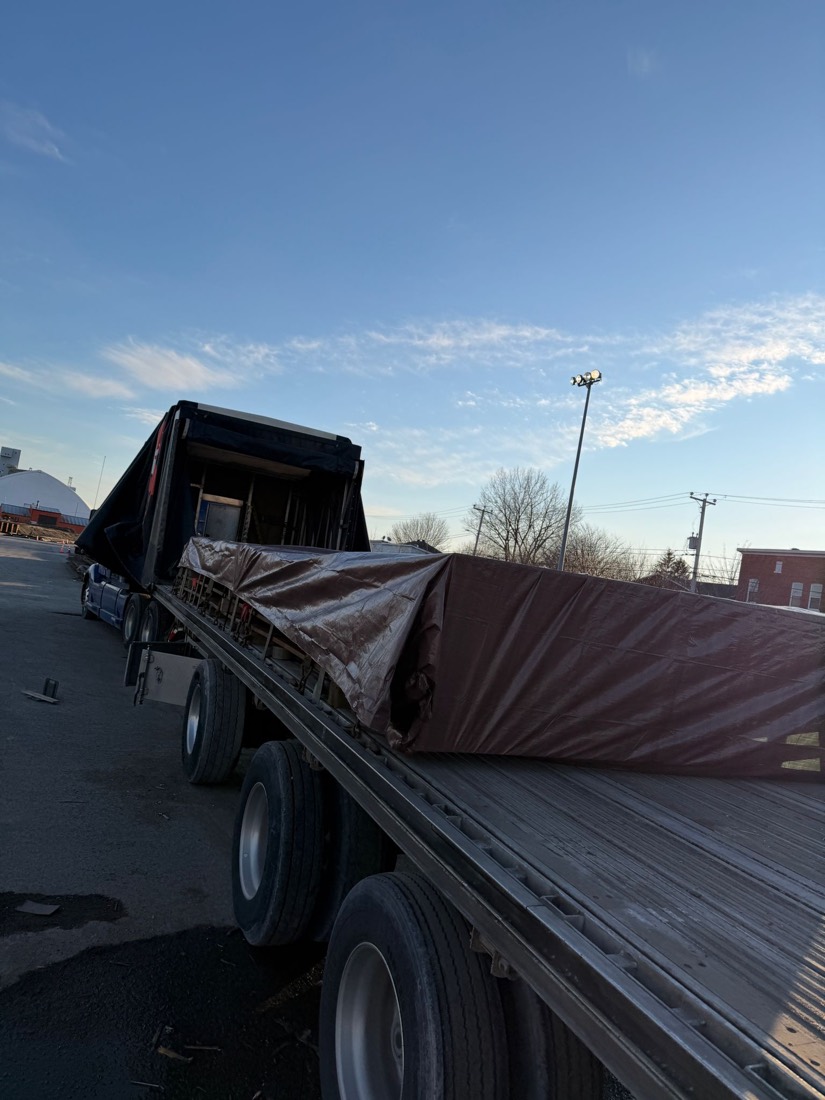 Brown-tarped load on curtain-frame flatdeck at golden hour, light snow on ground